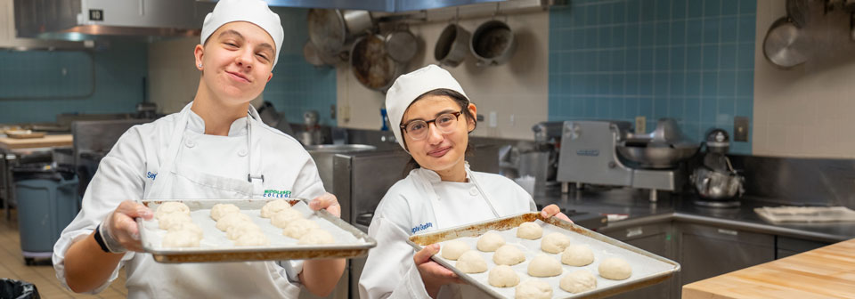 Two culinary arts students holding trays of baked goods.