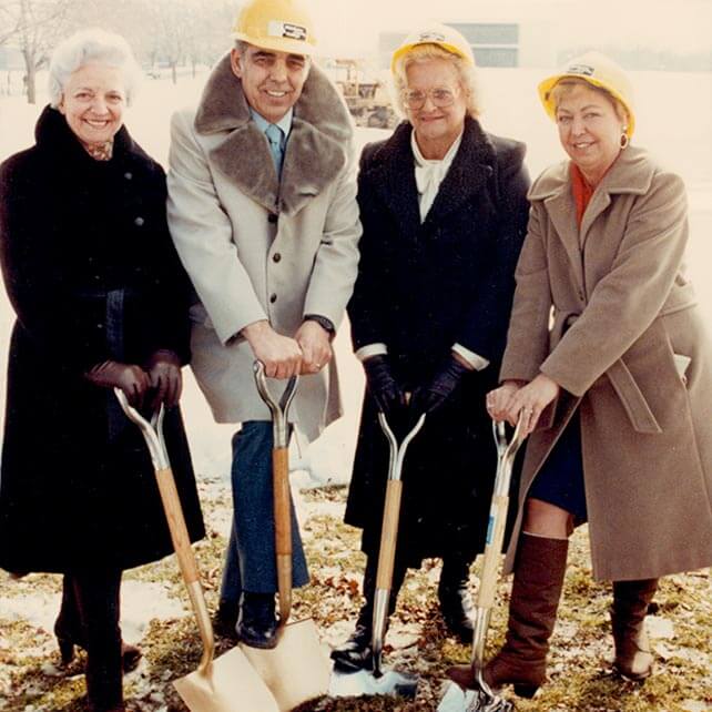 Dot Powers, shovel-in-hand alongside three others, gathers for a ground breaking for a new building. Patches of snow cover the grass.