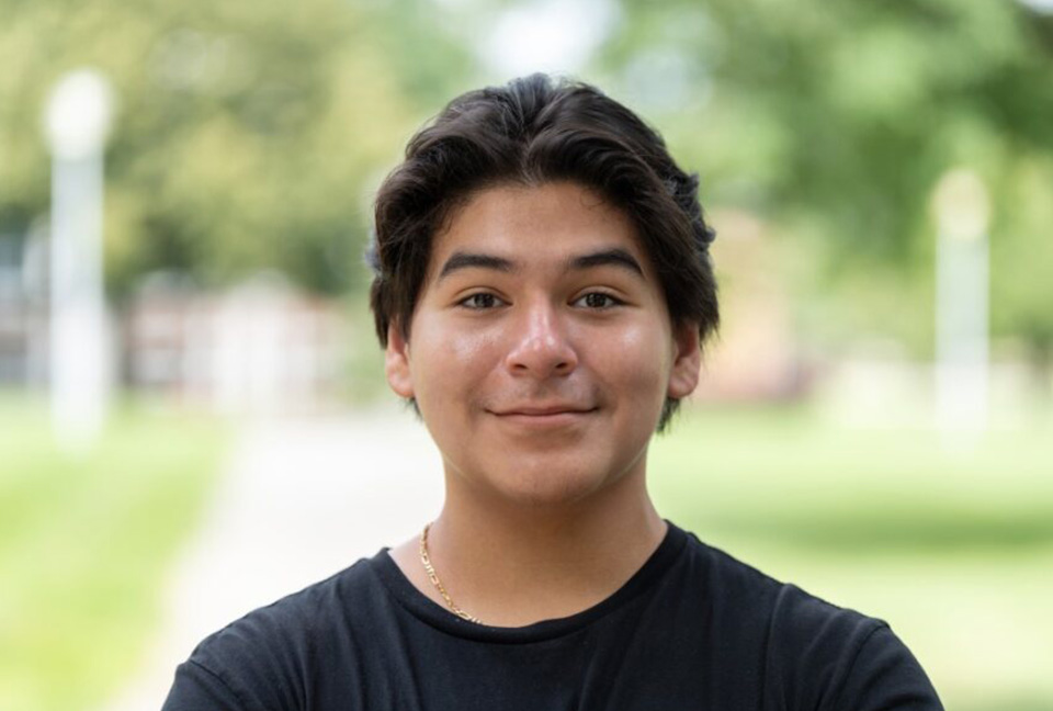 Gustavo, wearing a blank shirt and gold chain, smiles for a photo outside on the Edison campus.