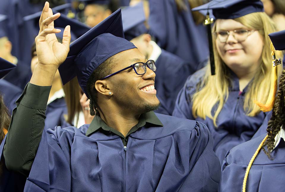 Close-up of a male graduate who is seated and raising his arm as he looks out to people in the stands.