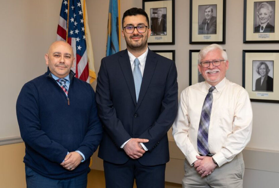 Jack Kent Cooke Scholarship winner Nima Sedghi is flanked by José Laureano, Vice President, Student & Enrollment Services, and Middlesex College President Mark McCormick, with the American flag and black and white photos behind them.