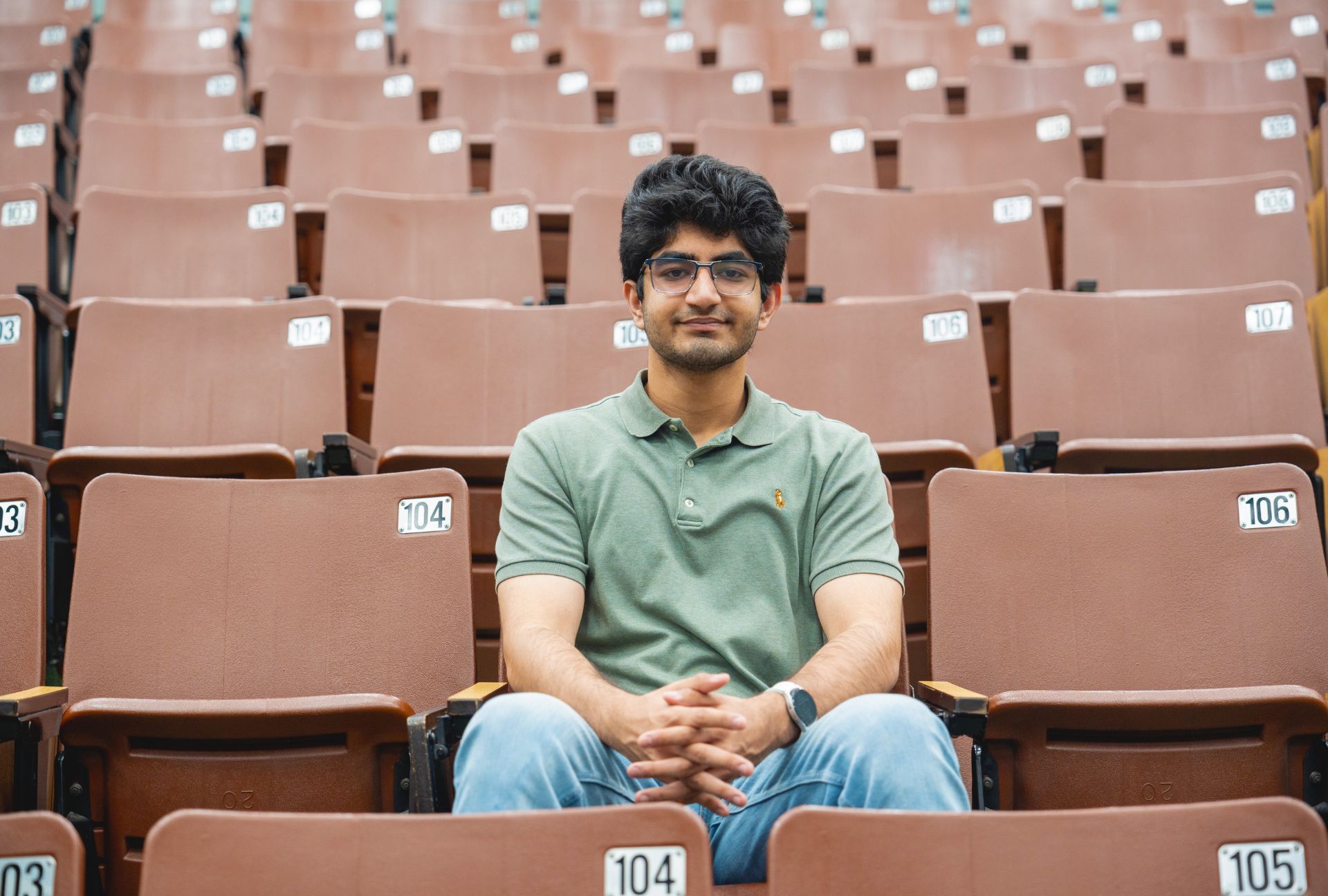 Dhruv Sethi, '25 Salutatorian, sitting with hands folded in a room of tiered seats.