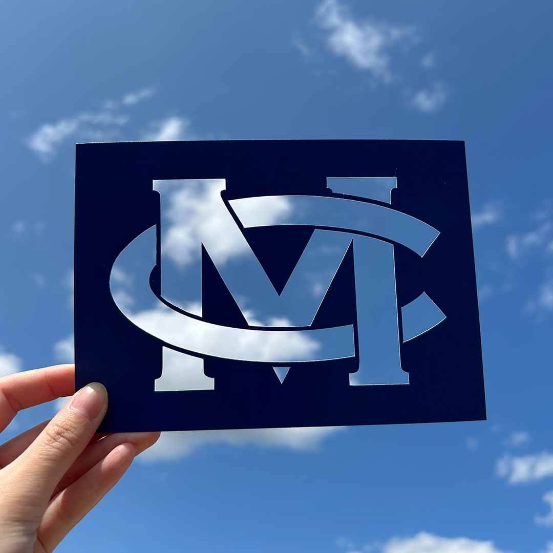 An 'MC' cutout held up to a blue sky with clouds.