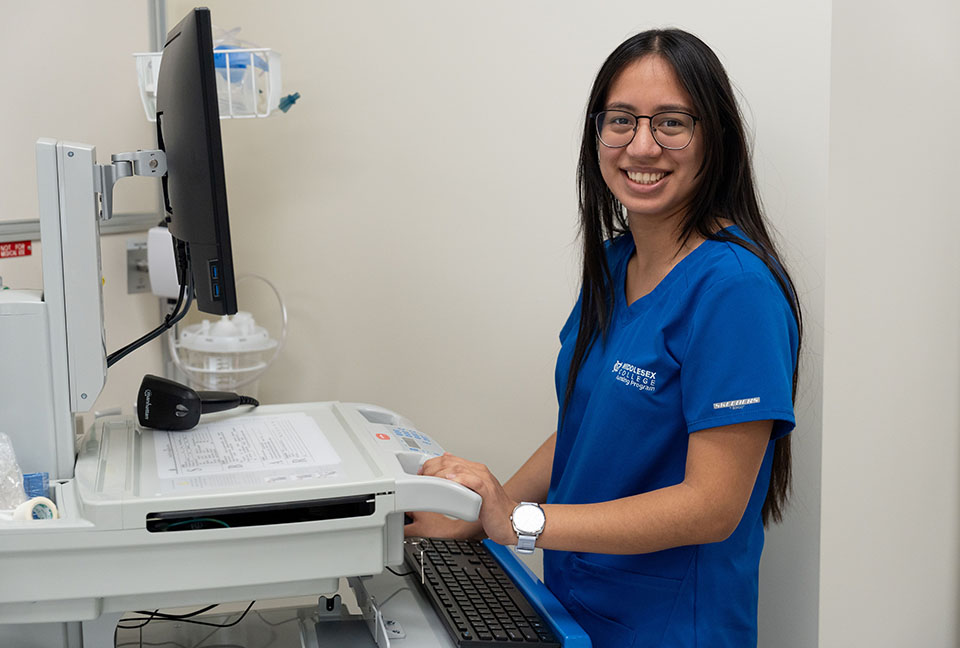 Nursing student in blue scrubs stands at a computer screen in office setting